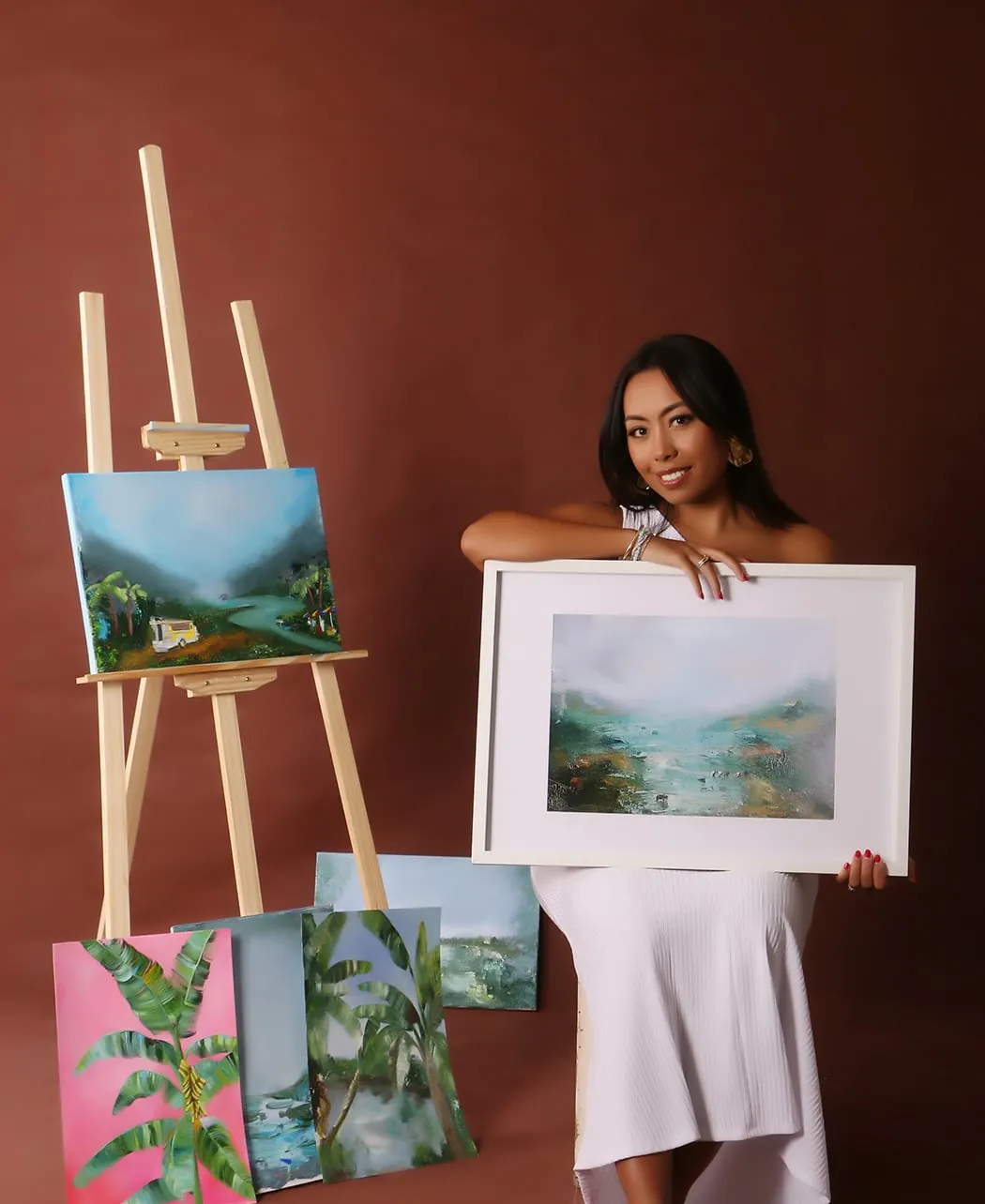 Margherita holding a framed landscape beside her easel
